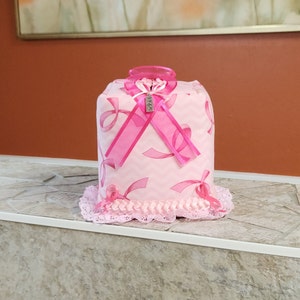 May include: Pink tissue box cover with a pink ribbon design and a charm that says "Hope". The cover has a lace trim and is sitting on a tiled surface.