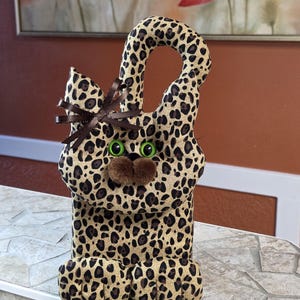 May include: A stuffed animal cat-shaped bag with a leopard print pattern. The bag has green eyes, a brown nose, and a brown bow. The bag is sitting on a white and gray surface.