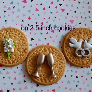 May include: Three round cookies, each about 2.5 inches in diameter, decorated with wedding-themed designs. One cookie features a white flower bouquet, another has two champagne glasses, and the last displays two white doves with silver rings.
