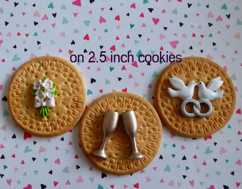 May include: Three round cookies, each about 2.5 inches in diameter, decorated with wedding-themed designs. One cookie features a white flower bouquet, another has two champagne glasses, and the last displays two white doves with silver rings.