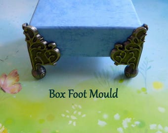 May include: Close-up of a light blue box with ornate, bronze-colored corner feet. The feet have a decorative, scrolled design and a rounded base. The text "Box Foot Mould" is visible in the image.