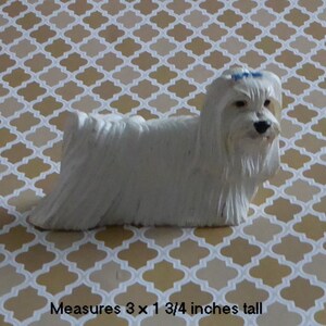 May include: A white Maltese dog figurine with a blue bow. The figurine measures 3 x 1 3/4 inches. The dog has long, flowing fur and is posed in a standing position. The background is a patterned design in shades of beige and white.