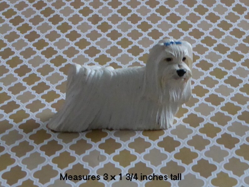 May include: A white Maltese dog figurine with a blue bow. The figurine measures 3 x 1 3/4 inches. The dog has long, flowing fur and is posed in a standing position. The background is a patterned design in shades of beige and white.