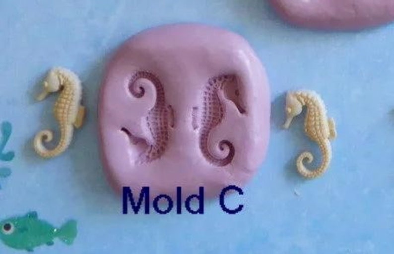 K&ouml;nnte beinhalten: Auf hellblauem Hintergrund befindet sich eine rosa Form mit zwei Seepferdchen-Abdr&uuml;cken und zwei separaten Seepferdchen-Figuren. Der Text "Mold C" ist blau gedruckt. Ein gr&uuml;ner Fisch ist ebenfalls zu sehen.