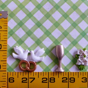 May include: A collection of wedding-themed miniatures. Includes two white doves kissing above two gold rings, a champagne flute, and a small bouquet of white flowers with a bow. The items are arranged on a green and white checkered background.