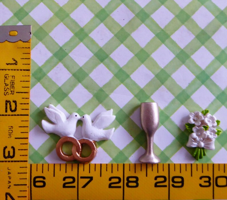 May include: A collection of wedding-themed miniatures. Includes two white doves kissing above two gold rings, a champagne flute, and a small bouquet of white flowers with a bow. The items are arranged on a green and white checkered background.
