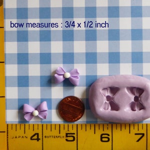 May include: Two small, light purple bow-shaped embellishments with white centers, next to a penny and a lavender mold. The text "bow measures: 3/4 x 1/2 inch" is visible. The background is a blue and white checkered pattern.
