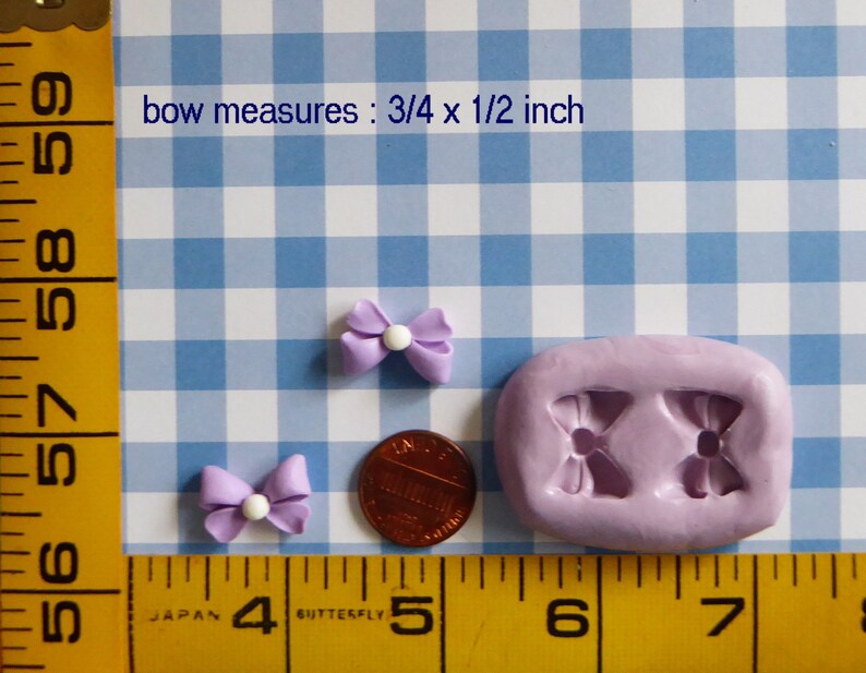 May include: Two small, light purple bow-shaped embellishments with white centers, next to a penny and a lavender mold. The text "bow measures: 3/4 x 1/2 inch" is visible. The background is a blue and white checkered pattern.