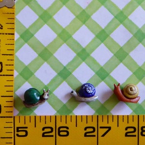 May include: Three miniature snail figurines. One has a green shell, another a blue shell with white accents, and the last a yellow shell. The snails are on a green and white checkered background. The image includes a yellow ruler.