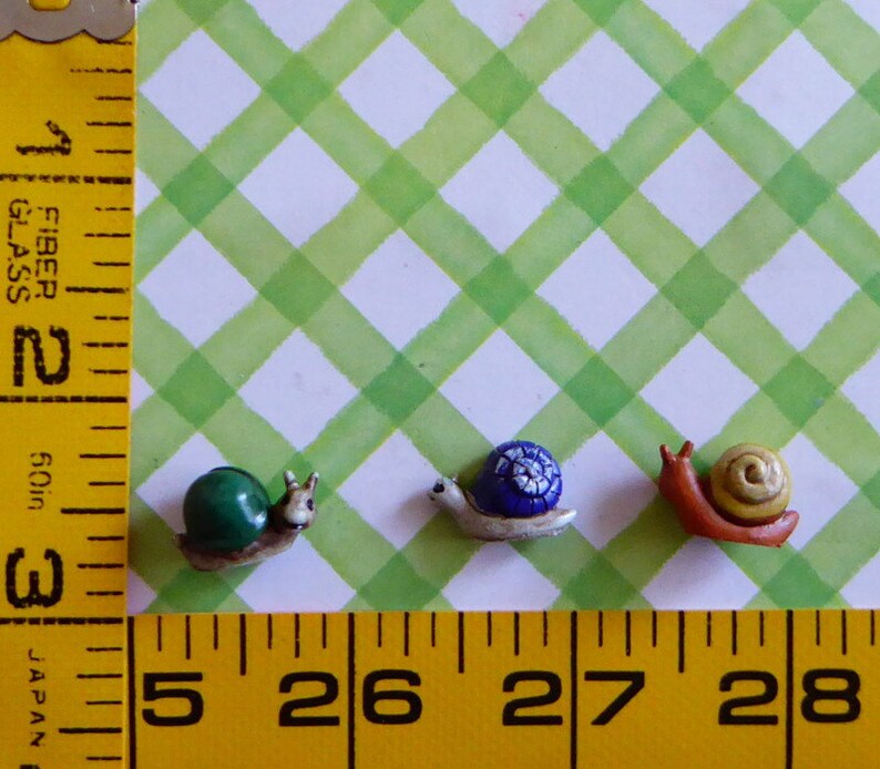 May include: Three miniature snail figurines. One has a green shell, another a blue shell with white accents, and the last a yellow shell. The snails are on a green and white checkered background. The image includes a yellow ruler.