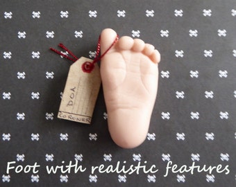 May include: A lifelike, pale pink foot with detailed features, accompanied by a small, brown tag. The tag is attached with red string and has the words "DOA" and "CORONER" written on it. The background is black with a white skull pattern.