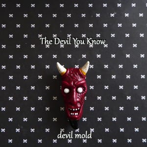 Könnte beinhalten: Eine rote Teufelsform mit weißen Augen und Hörnern sowie einem schwarzen Kinn. Der Hintergrund ist schwarz mit einem Muster aus weißen Totenköpfen. Der Text "The Devil You Know" und "devil mold" sind sichtbar. Die Teufelsform hat ein breites Grinsen.