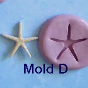 K&ouml;nnte beinhalten: Ein Seestern und eine rosa Form mit einem Seestern-Abdruck werden auf einer hellblauen Oberfl&auml;che gezeigt. Der Text "Mold D" ist darunter in Dunkelblau geschrieben. Der Seestern ist beige.