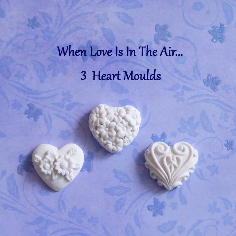 May include: Three white heart-shaped molds with different floral and decorative designs. The molds are arranged on a light purple background with a floral pattern. Text at the top reads "When Love Is In The Air... 3 Heart Moulds."