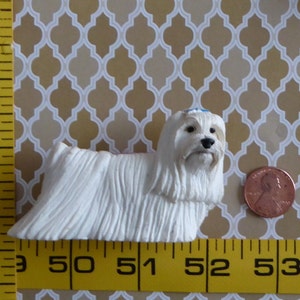 May include: A small, white Maltese dog figurine with long, flowing fur. The dog has a black nose and eyes, and a small blue bow. A penny is next to the dog for size comparison. The background is a patterned design.