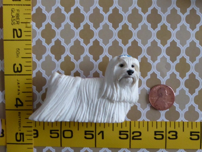 May include: A small, white Maltese dog figurine with long, flowing fur. The dog has a black nose and eyes, and a small blue bow. A penny is next to the dog for size comparison. The background is a patterned design.