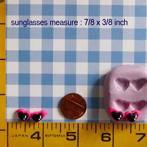 May include: Miniature heart-shaped sunglasses with black lenses and pink frames. The sunglasses measure 7/8 x 3/8 inch. A penny and a mold with heart-shaped sunglasses are also in the image. The background is a blue and white checkered pattern.