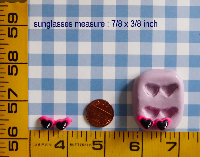 May include: Miniature heart-shaped sunglasses with black lenses and pink frames. The sunglasses measure 7/8 x 3/8 inch. A penny and a mold with heart-shaped sunglasses are also in the image. The background is a blue and white checkered pattern.