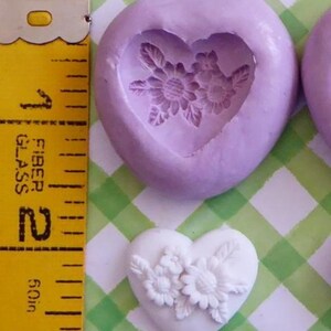May include: A heart-shaped mold with a floral design, next to a finished white heart with flowers. The mold is lavender, and the finished heart is white. A yellow measuring tape shows the size of the objects.