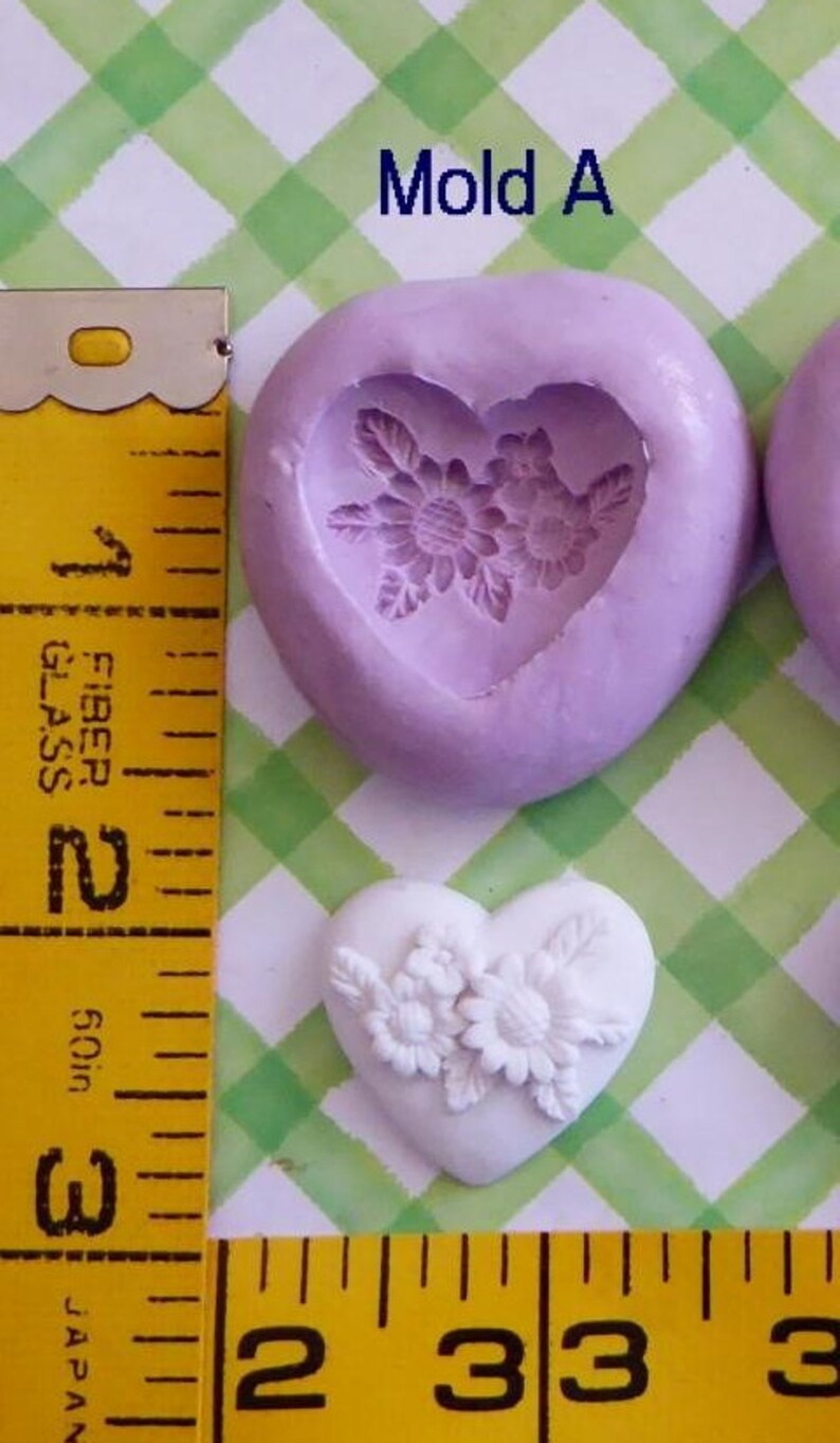 May include: A heart-shaped mold with a floral design, next to a finished white heart with flowers. The mold is lavender, and the finished heart is white. A yellow measuring tape shows the size of the objects.