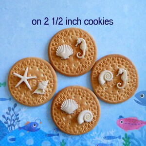 K&ouml;nnte beinhalten: Vier runde Kekse, jeweils etwa 6,35 cm im Durchmesser, verziert mit wei&szlig;en Muschel- und Seepferdchen-Designs. Ein Keks hat einen Seestern. Der Text "on 2 1/2 inch cookies" ist sichtbar.