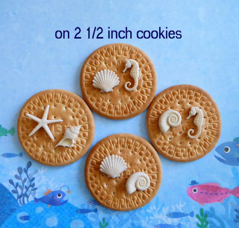 K&ouml;nnte beinhalten: Vier runde Kekse, jeweils etwa 6,35 cm im Durchmesser, verziert mit wei&szlig;en Muschel- und Seepferdchen-Designs. Ein Keks hat einen Seestern. Der Text "on 2 1/2 inch cookies" ist sichtbar.