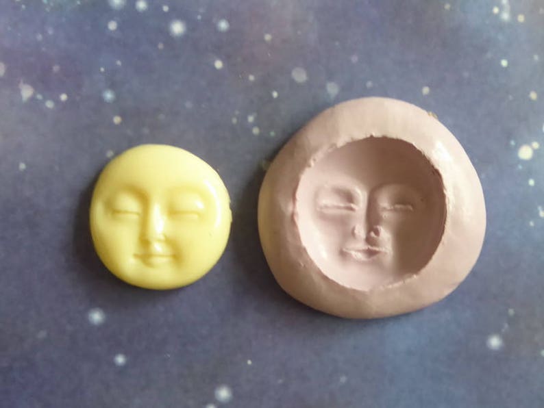 May include: A yellow, circular moon face with closed eyes sits next to a pink, oval-shaped mold of a moon face. The moon face has a serene expression. The background is a dark blue with white speckles.
