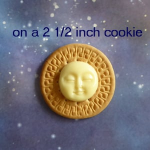 May include: A round, tan cookie with a cream-colored sun-shaped decoration. The cookie is approximately 2.5 inches in diameter. The text "on a 2 1/2 inch cookie" is written above the cookie against a blue background.
