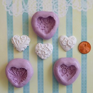 May include: An assortment of heart-shaped molds and decorative heart embellishments. The molds are lavender, with floral and ornate designs. The embellishments are white, featuring floral and scrollwork patterns. A penny is included for size reference.