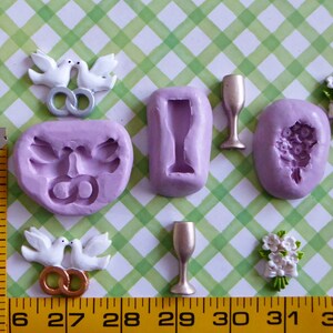 May include: A collection of wedding-themed craft supplies, including molds for doves, champagne glasses, and a bouquet, along with finished dove and bouquet embellishments. The items are in shades of white, gold, and lavender, arranged on a green and white checkered surface.