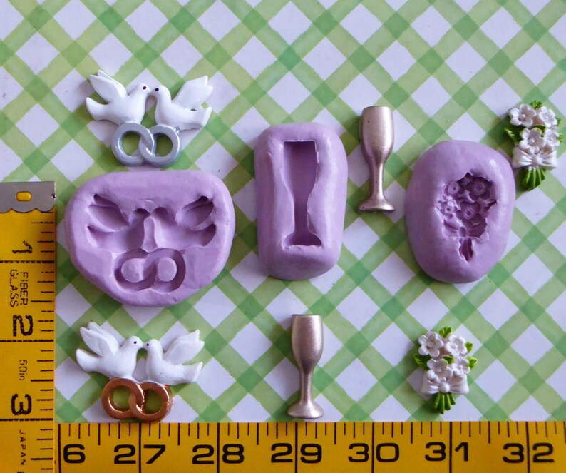 May include: A collection of wedding-themed craft supplies, including molds for doves, champagne glasses, and a bouquet, along with finished dove and bouquet embellishments. The items are in shades of white, gold, and lavender, arranged on a green and white checkered surface.
