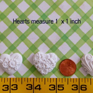 May include: Three white heart-shaped embellishments with different floral and scroll designs. The hearts measure 1 x 1 inch. A penny is next to the hearts for size comparison. The background is a green and white checkered pattern.