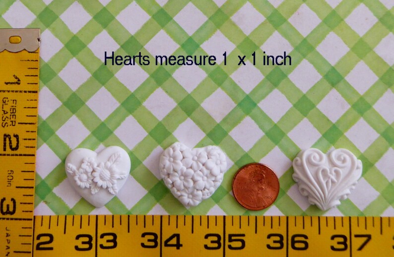 May include: Three white heart-shaped embellishments with different floral and scroll designs. The hearts measure 1 x 1 inch. A penny is next to the hearts for size comparison. The background is a green and white checkered pattern.