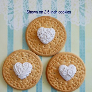 May include: Three round cookies, each with a white heart-shaped decoration. The cookies are approximately 2.5 inches in diameter. The decorations feature floral and ornate designs. The background is a light blue and yellow striped pattern.