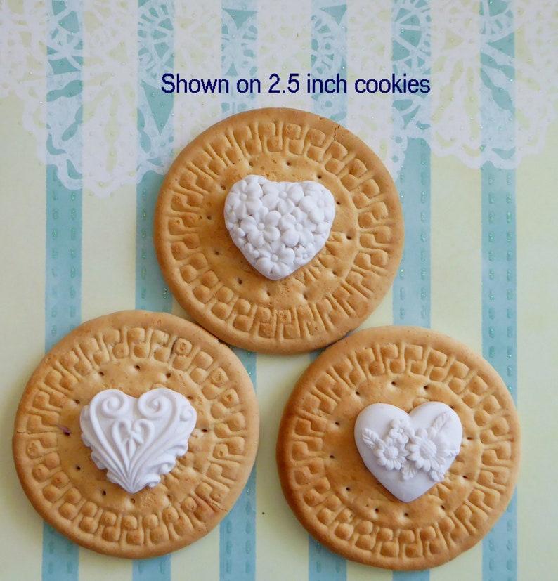 May include: Three round cookies, each with a white heart-shaped decoration. The cookies are approximately 2.5 inches in diameter. The decorations feature floral and ornate designs. The background is a light blue and yellow striped pattern.