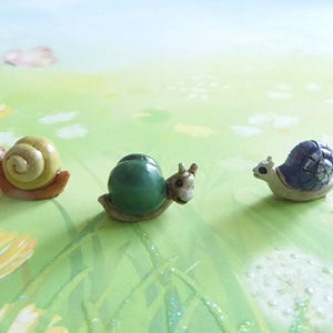 May include: Three miniature snail figurines with detailed shells in shades of yellow, green, and blue. The snails are set against a backdrop of a green and yellow meadow with dandelions and a light blue sky.