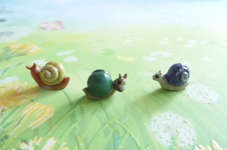 May include: Three miniature snail figurines with detailed shells in shades of yellow, green, and blue. The snails are set against a backdrop of a green and yellow meadow with dandelions and a light blue sky.