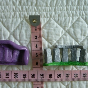 May include: Miniature purple object and a Stonehenge replica next to a penny, with a pink measuring tape showing inches. The purple object is approximately 1.5 inches long. The Stonehenge replica is about 1 inch wide.