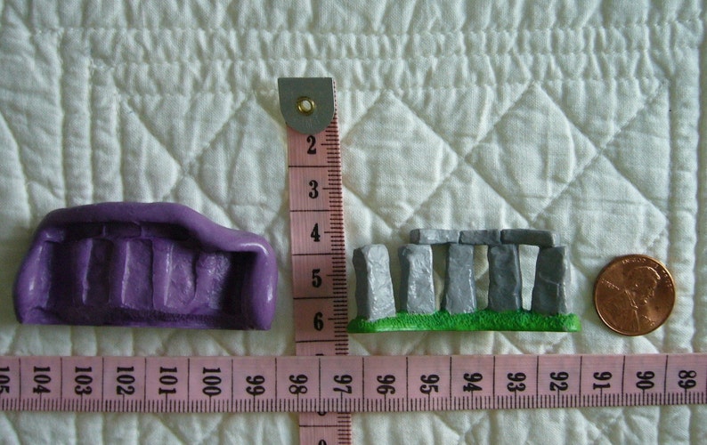 May include: Miniature purple object and a Stonehenge replica next to a penny, with a pink measuring tape showing inches. The purple object is approximately 1.5 inches long. The Stonehenge replica is about 1 inch wide.