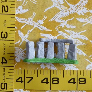 May include: A miniature Stonehenge replica with gray stone pillars and lintels, set on a green base. The model is displayed against a yellow background with a white bird pattern. A yellow measuring tape is in the foreground.