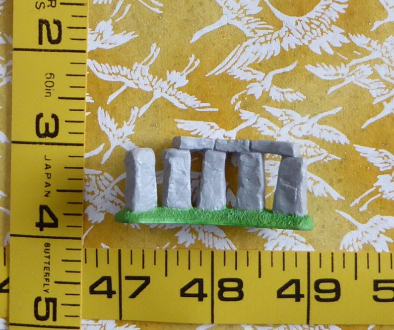 May include: A miniature Stonehenge replica with gray stone pillars and lintels, set on a green base. The model is displayed against a yellow background with a white bird pattern. A yellow measuring tape is in the foreground.
