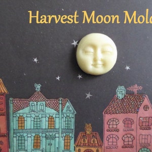 May include: A cream-colored Harvest Moon Mold with a smiling face, set against a dark background with a starry night sky and colorful illustrated houses. The text "Harvest Moon Mold" is in gold.