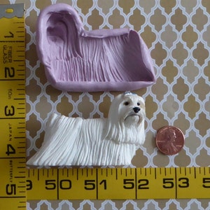May include: A white dog figurine with long, textured fur, next to a penny and a lavender mold. The dog is approximately 3 inches long. The mold is a light purple color. The background is a gold and white patterned surface.