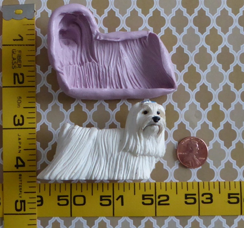May include: A white dog figurine with long, textured fur, next to a penny and a lavender mold. The dog is approximately 3 inches long. The mold is a light purple color. The background is a gold and white patterned surface.