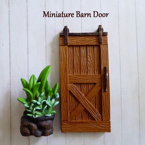 May include: A miniature brown barn door with a black handle and hardware, next to a small green plant in a brown pot. The door has an X-shaped design and the text "Miniature Barn Door".