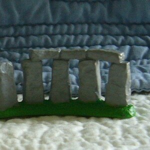 May include: A miniature Stonehenge replica. The model features gray stone structures atop a green base, set against a textured background. The iconic prehistoric monument is depicted in a small-scale, decorative form.