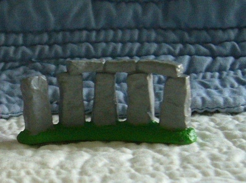 May include: A miniature Stonehenge replica. The model features gray stone structures atop a green base, set against a textured background. The iconic prehistoric monument is depicted in a small-scale, decorative form.