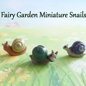 May include: Three miniature fairy garden snails. One snail has a brown body and a tan shell, another has a green shell, and the third has a blue shell. The text "Fairy Garden Miniature Snails" is at the top of the image.