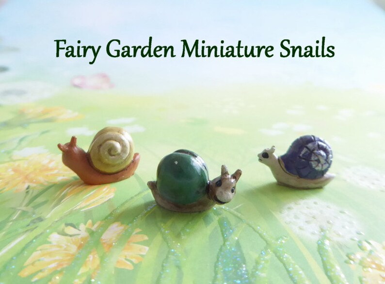 May include: Three miniature fairy garden snails. One snail has a brown body and a tan shell, another has a green shell, and the third has a blue shell. The text "Fairy Garden Miniature Snails" is at the top of the image.