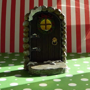 May include: A miniature fairy door with a dark wooden door, stone frame, and a small wreath. The door has a brass door knocker and is decorated with faux snow. The background features a red and white striped pattern.